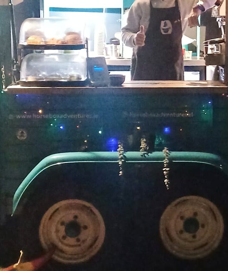 The hatch counter of the Horsebox Adventures cart at night: pastries in the display cases, a card reader and the wheel arch below, with fairy lights along the side panel.