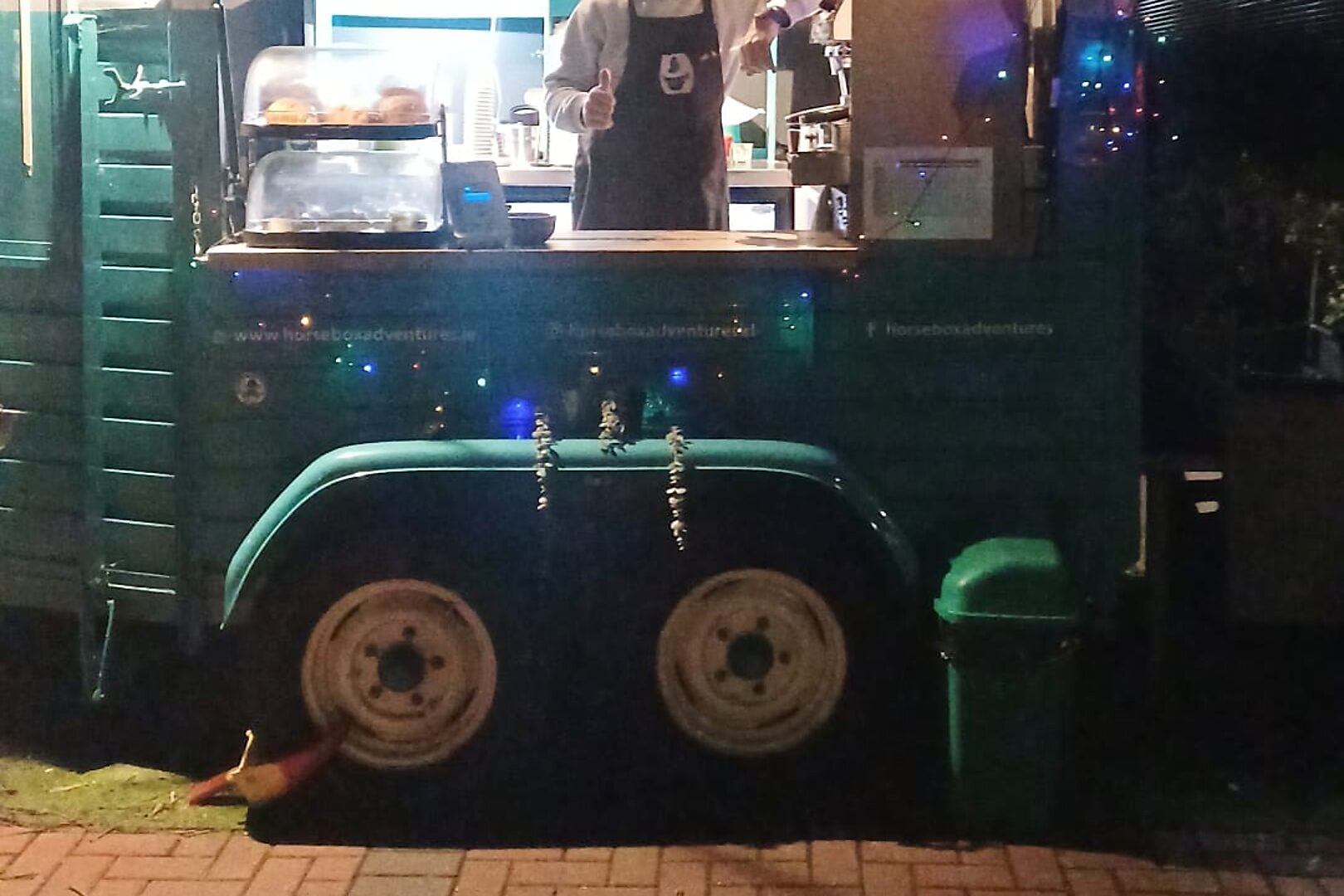 The Horsebox Adventures cart at night, lit by fairy lights, with pastries in the display on the hatch counter and the shop's web and social handles painted on the teal-green side panel.